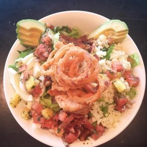 A plate full of rice, tomatoes, lettuce, avocado, onion rings and bacon